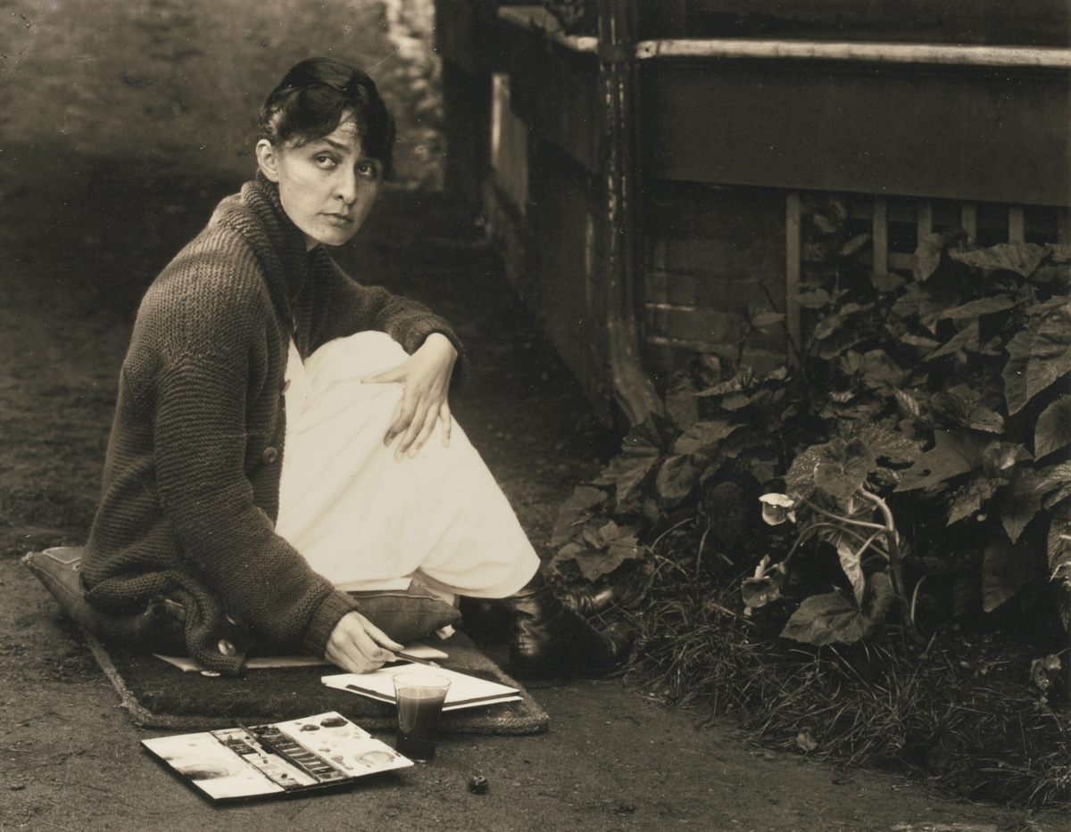 Alfred Stieflitz, Photograph of Georgia O&#x27;Keeffe, seen nestled on a cushion on the ground, with her sketchpad and watercolors by her side, 1918. Image via Wikimedia Commons. 