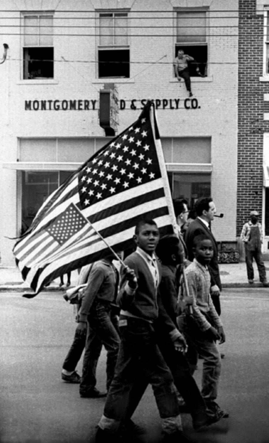 Young boys marching with American Flags past Montgomery Feed & Supply Co., with man perched on upper floor window sill, March 25, 1965