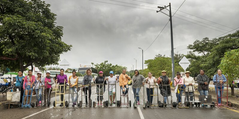 Teresa Margolles, ‘Carretilleras sobre el puente internacional Simon Bolivar (Porters on the international bridge Simon Bolivar)’, 2018, Photography, The Venezuelan crisis has generated an exodus towards neighboring countries. On the bridge that crosses the Táchira River connecting the cities of Cúcuta and San Antonio, the occupation of the so-called „Carretillero“ is thriving. The term describes people dedicated to carry goods acquired in Colombia to Venezuela by crossing the bridge. This occupation is carried out by Venezuelans and was first executed exclusively by men. In recent months, due to the lack of employment and the increasing socio-economic crisis, women have more and more taken this occupation as well. Focusing on this specific situation, the artist invited the respective women, who were operating in different parts of the area called „La Parada“ next to the Simon Bolivar Bridge, to carry out an action that consisted of lining their working equipment  - the wheelbarrow - to form a line on the bridge. The action was done on 3 days in a row and lasted 3 minutes. Cúcuta, Colombia Color print, Galerie Peter Kilchmann