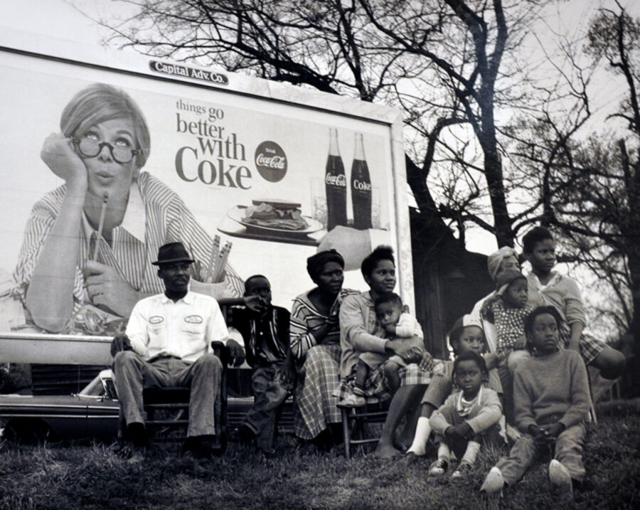 Coca Cola lady looking over black multi-generational family seated on hillside under sign, watching 1965 Selma to Montgomery civil march pass by, March 25, 1965