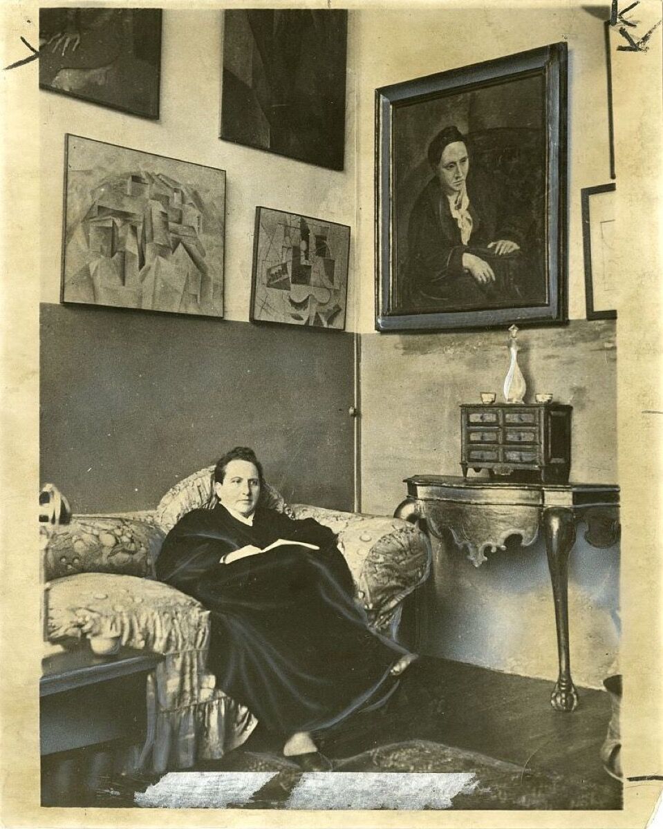 Gertrude Stein sitting on a sofa in her Paris studio, with a portrait of her by Pablo Picasso, and other modern art paintings hanging on the wall behind her, 1930. Image via U.S. Library of Congress.
