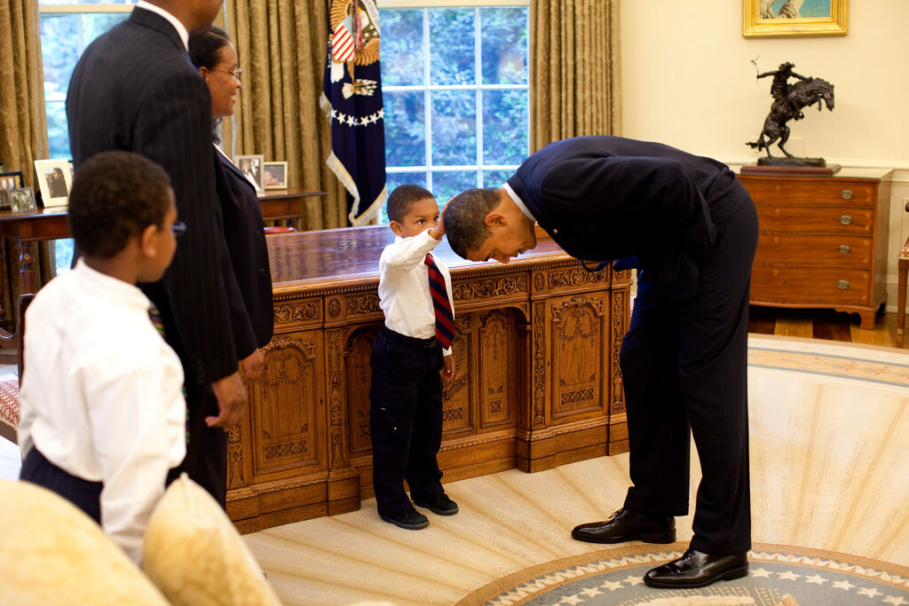 President Barack Obama bends over so the son of a White House staff member can pat his head during a family visit to the Oval Office, 2009. Photo by Pete Souza. Courtesy of the White House Photo Office via Flickr.